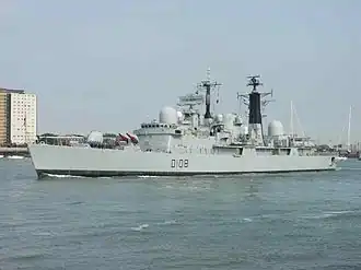 Grey warship with black towers and red missiles on its bow, city buildings are in the background.