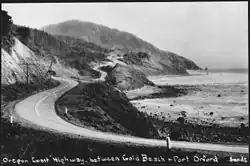 Black-and-white photograph of a narrow road winding around the cliffs overlooking the ocean.
