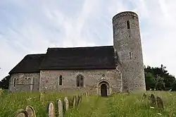 A stone church with thatched roofs seen from the north; on the left is a chancel with an apse, in the middle is the nave containing a Norman doorway, and on the right is a round tower