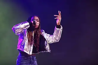 Haviah Mighty wearing dark blue jeans and a shiny silver jacket, singing into a microphone onstage