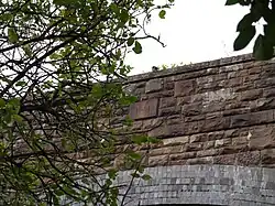 The bridge from the road, looking up through foliage at the east parapet wall with its unornamented date stone.