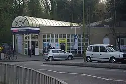 A single-storey yellow-brick building with its entrance at one of the corners. The entrance is covered by a glass canopy with a sign that reads "Hertford North". There are cars, bikes and people in front.