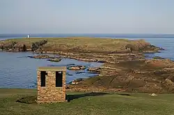 In the foreground is a building from the former RAF Skaw and in the background is the Holm of Skaw, with its lighthouse. At low tide it seems that it should be possible to walk out to the holm, but a deep channel runs between the rocks.