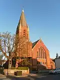 A dark red church dominated by a tower on the left. This has stone spirelets at each corner and a central slate-coloured spire, and is partly obscured by trees. There are five lancet windows in a slight recess in the wall to the right.