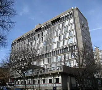 A tall concrete building seen from below and to the right, with a front facade consisting mainly of windows. Two small bare trees are in front.