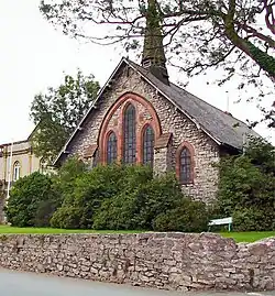 The end of a chapel with five windows and a small spire; in front is a bush and a wall