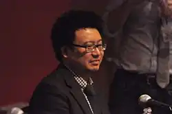 Hua Hsu sits at a table in front of a microphone wearing black glasses and a jacket and tie with a checkered shirt