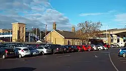 A small yellow-brick single-storey building with a slanted roof and chimneys viewed from some distance away .The view of the building is mainly blocked by a large row of cars. To the left is a visible blue footbridge.
