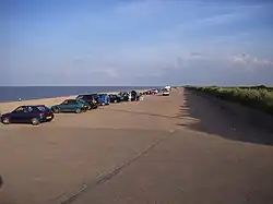 cars lined up stretching away along hard surfaced standing next to a golden sand beach and sea beyond against a blue sky on a sunny day