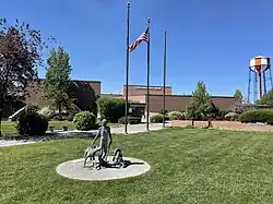 In front of a modern brick building sits a neatly manicured lawn featuring a sculpture of a person and two dogs, and three flagpoles with the American flag flying from one of them.
