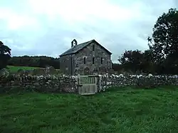 A small, simple, stone church, in front of which is a stone wall. On the far gable is a bellcote with a single bell.