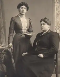 An image of two women. One is standing with a book in her left hand and is resting her right hand on a table. The other woman is seated in a wooden chair.