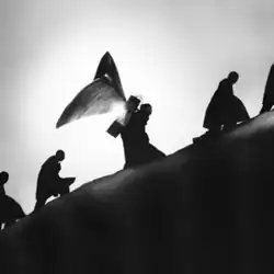 A black-and-white silhouetted photo of multiple people walking up a steep slope, with one in the middle carrying a butterfly-shaped object on their back.