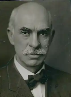 A black and white photo of a bald white man with a bushy mustache and a bowtie gazing sternly at the camera.