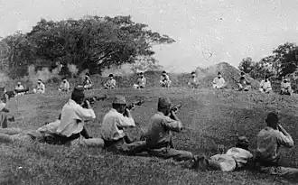 A dozen prisoners sit bound at a distance as Japanese soldiers in the foreground kill them