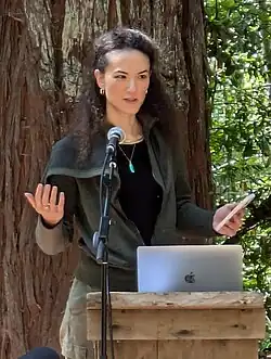 Portrait of Jay Graber, a Chinese–American woman with long curly hair, wearing casual clothing. She is giving a talk while outside, holding a phone behind a podium. There is a microphone on a stand and a laptop in front of her.