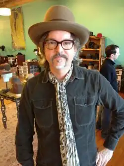 A man with a goatee stands in a room with his left hand on his hip. He is wearing a brown fedora, dark-brown glasses, a pocketed dark-teal shirt, and a leopard-print scarf.