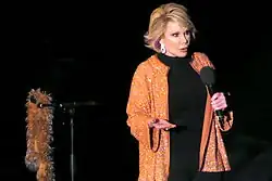 A woman with short blond hair wearing black except for a sparkly orange jacket with earrings. She is holding a microphone, and next to her is a microphone stand with an orange boa hanging from it.