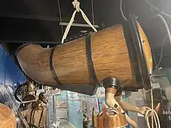 Tapered one-person submarine like wooden vessel with sealed holes for operator's arms held aloft by rope slings in a museum display