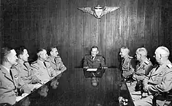 Black and white photograph. A man in dark khakis is at the head of the table, with three men seated to his left side and four to his right