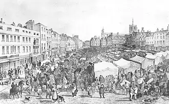 Very crowded market on a slope. At the left of the picture is a row of tall thin shops, and behind the market is another row of shops surrounding a tall church.