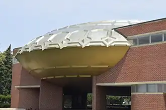 View of the Golden Rondelle Theater's facade following its relocation to the Johnson Wax Headquarters. The roof is a gold-colored disk with embossed shapes. There are brick pavilions on either side of the disk.