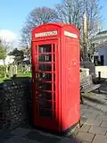 Three-quarter view of a red telephone booth with a glass door with red metal bars (partly in shadow) and a slightly domed roof. Behind it stand a flint wall, gravestones, a wooden bench and a tall stone war memorial.