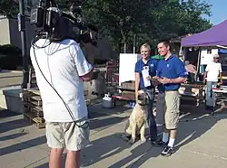 Two employees in KGAN polo shirts (with the CBS 2 logo on the chest and the Fox 28 logo on the sleeve), filmed by a KGAN cameraman, at an event