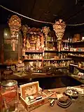 Interior photograph of the Kam Wah Chung Company building showing a general store area, dimly lit, with many products on counters, tables, shelves, and hanging from the wood plank ceiling. Many products are conspicuously Chinese in style, and all furniture and building elements are wooden.