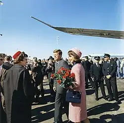 Kennedy and the First Lady, dressing in a pink outfit and holding a bouquet of flowers, depart from Air Force One and greet welcomers