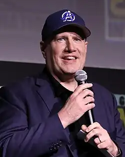 Kevin Feige speaking at the 2014 San Diego Comic-Con wearing a suit and a hat with a purple Avengers insignia.