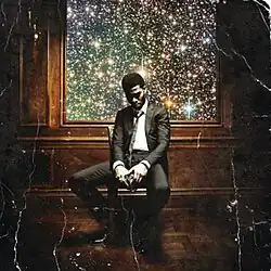 Kid Cudi sitting in a chair with fireworks in a window in the background.