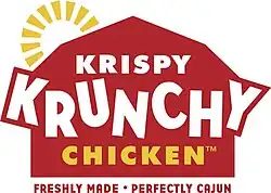 The text "Krispy Krunchy Chicken" in white and yellow letters, on a stylized depiction of a red barn with a yellow sun behind it. Below the barn is the text "Freshly made. Perfectly Cajun." in red letters.