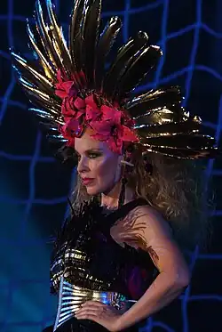 An image of Kylie Minogue holding her waist and wearing a black dress, silver belt and a headpiece with red and gold details.