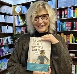 Color photo of a woman holding a book, the bookshelves behind her filled with other books.