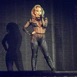 Lady Gaga standing on a stage in a mesh leather cut-out bodysuit and knee-high leather boots. Half of her hair is dyed green.