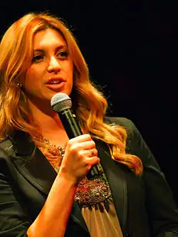 A photograph of a blonde woman speaking into a black microphone which she is holding in her right hand while wearing a black jacket
