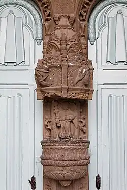 The highly decorated stoup in the porch of Landivisiau's Église Saint-Thuriau. This is fixed to the trumeau between the two entrance doors to the church. It is decorated with godrons (gadrooning) and beneath an elaborate canopy is the depiction of an angel holding a bottle brush or aspergillum ("Goupillon"), an allusion to the baptism. The renaissance dais features flat pilasters decorated with acanthus leaves, seashells and fire pots and the cartouches below depict four high relief carvings; the heads of two men, their feathered hats "à la Henri 111" alternating with the heads of two women whose long hair is held back by a hairband in the Renaissance style.
