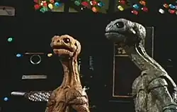 Two reptile-like alien creatures, one blue and one orange, look in an upward direction, with machinery and computer screens in the background.