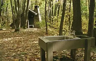 A metal sink with running water supported by wooden beams in the foreground, a wooden open shelter with a stone chimney amidst the trees in the background