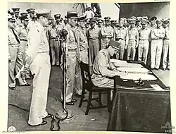 A large crowd of men in uniforms. Leclerc sits at a small desk signing a document. MacArthur stands behind him.