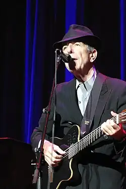 Leonard Cohen wearing a black fedora and a black suit. He is playing the guitar and singing into a microphone.