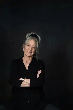 A woman in her mid 70s stands with her arms crossed and smiling. She is wearing all black and is against a black backdrop.