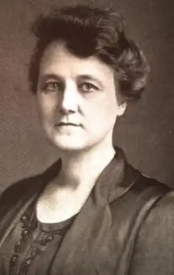 A middle-aged white woman, dark hair dressed off her shoulders, wearing a dark jacket and matching blouse or dress