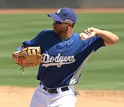 A man in a blue baseball jersey with "DODGERS" on the chest with a baseball glove on his right hand prepares to throw a baseball with his left hand.