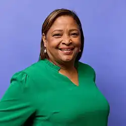 A photograph of Lori Montenegro, posed, facing the camera with a blue background. Montenegro is smiling and wearing a green blouse.