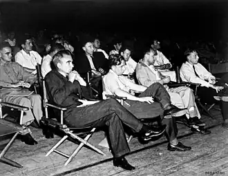 A group of formally dressed people sit in the audience, on folding chairs, and listen to a lecture