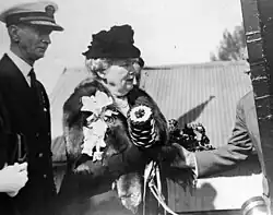 Black and white photograph of a men in a naval uniform jacket and white cap at the left, a women with a fur coat and hat at the centre