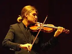 A man with a ponytail and black suit, looking to the right with a violin resting on his shoulder