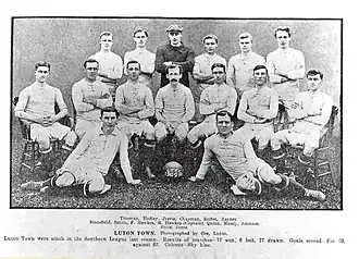 An association football team. Six men stand across the back, the third from the left of which is wearing a dark shirt and a cap. In front of them sits a row of seven men on chairs, with a mustachioed gentleman with hair that is quite long for the time sitting in the middle. Finally, two men recline on the floor in front of them, one on either side of a football marked "LTFC 1909–10".